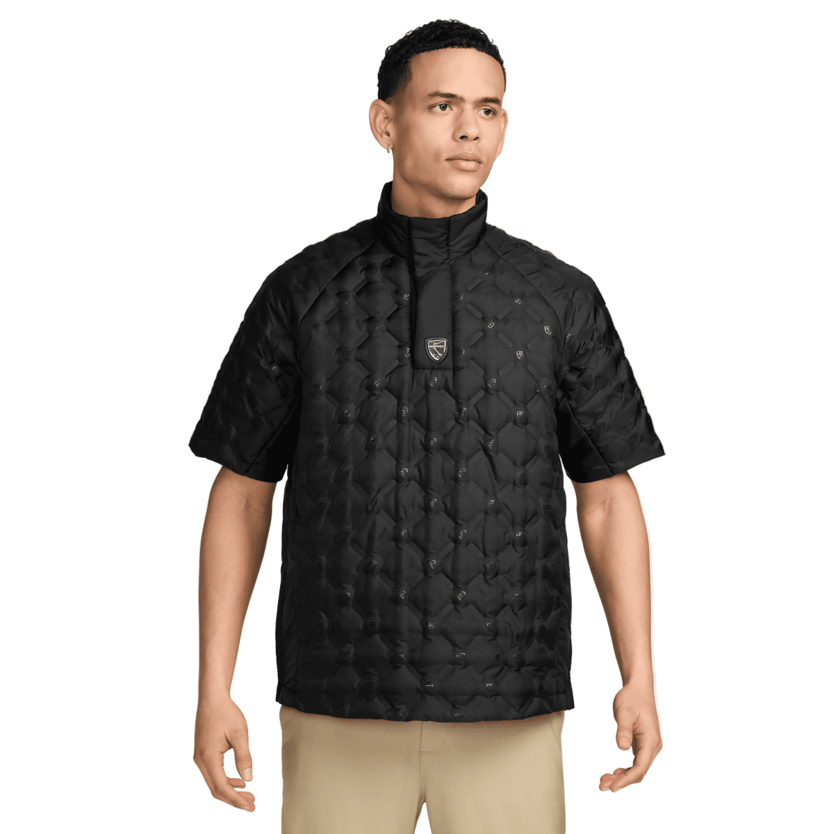 A man wearing a black short-sleeve quilted jacket with a stand-up collar and a small shield logo on the chest, paired with beige pants, against a light gray studio background.