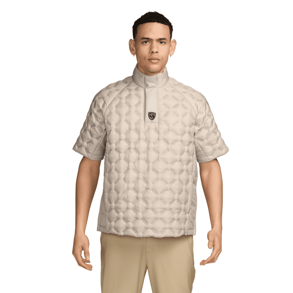 A man wearing a light beige short-sleeve quilted jacket with a stand-up collar and a small black shield logo on the chest, paired with khaki pants, against a light gray studio background.